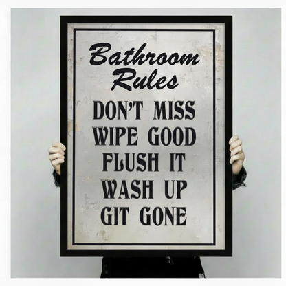 Person holding a large black framed Bathroom Rules humorous quote canvas art print showing print size scale and bold vintage distressed typography with Don't Miss Wipe Good Flush It Wash Up Git Gone text detail