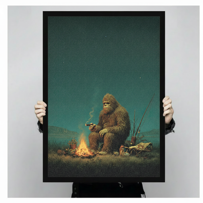 Person holding large framed retro realist oil painting Bigfoot sitting by campfire smoking cigar with fishing rods under starry night sky canvas print with both hands - close up view of humorous Sasquatch camping wall art with warm amber firelight deep teal sky and black frame finish