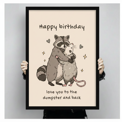 Hands holding a large black framed canvas print of a cartoon raccoon hugging an opossum with Happy Birthday lettering and sparkle details, funny animal birthday quote art on cream background against a light gray backdrop