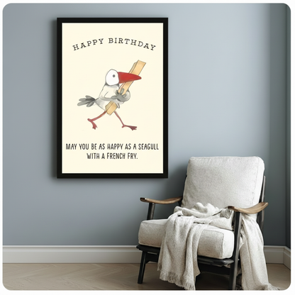 Black framed art print of a cartoon seagull strutting with a french fry and Happy Birthday lettering above, funny birthday quote canvas wall art on cream background, hung on a blue-gray wall beside a white upholstered armchair with a cream throw blanket