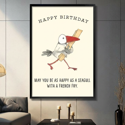 Black framed canvas wall art with Happy Birthday text and a cartoon seagull clutching a french fry, humorous hand-drawn illustration on cream background, displayed above a dark console table with pillar candles and black vase against a gray paneled wall