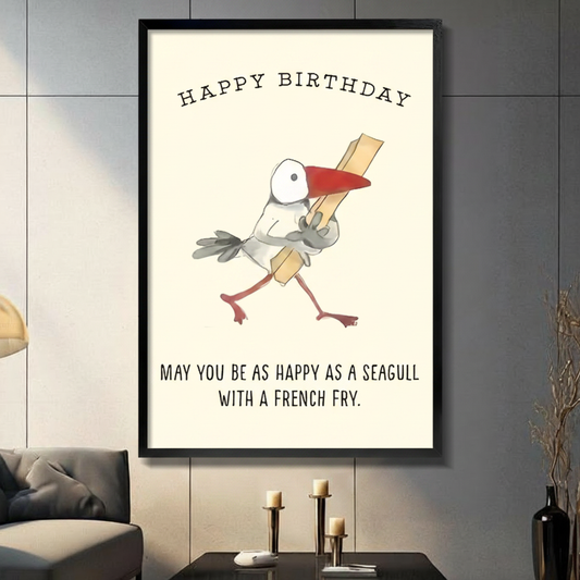 Black framed canvas wall art with Happy Birthday text and a cartoon seagull clutching a french fry, humorous hand-drawn illustration on cream background, displayed above a dark console table with pillar candles and black vase against a gray paneled wall