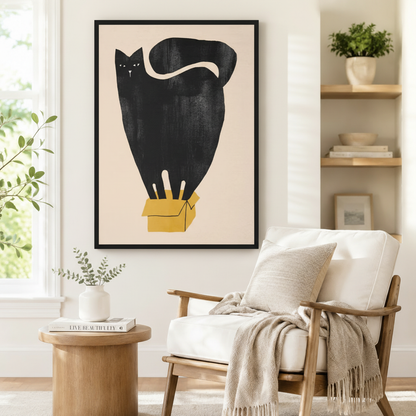Black framed abstract black cat sitting in mustard yellow cardboard box art print on white wall beside a white linen armchair and round wood side table