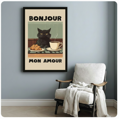 Black-framed funny cat wall art showing a grumpy fluffy black cat with yellow eyes beside a croissant and coffee cup with "Bonjour Mon Amour" text, styled on a soft blue-grey wall beside a white armchair