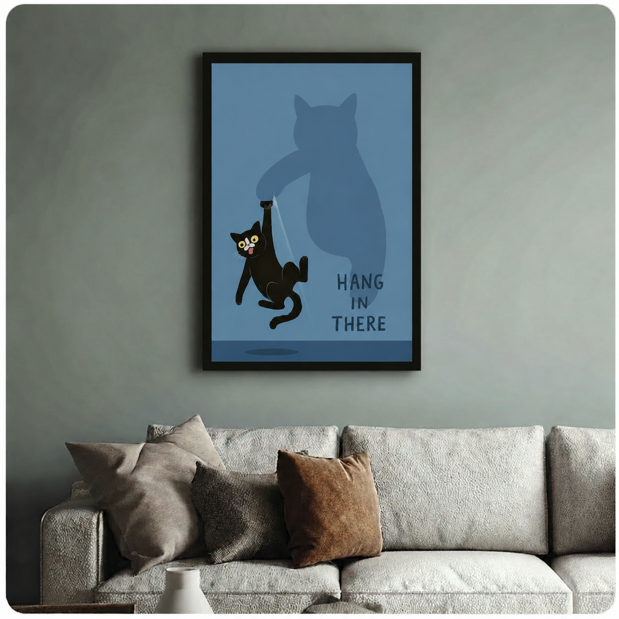 Cartoon black cat with shocked expression hanging from shadow cat's claw, bold blue-grey "Hang In There" motivational print, black framed wall art above beige fabric sofa