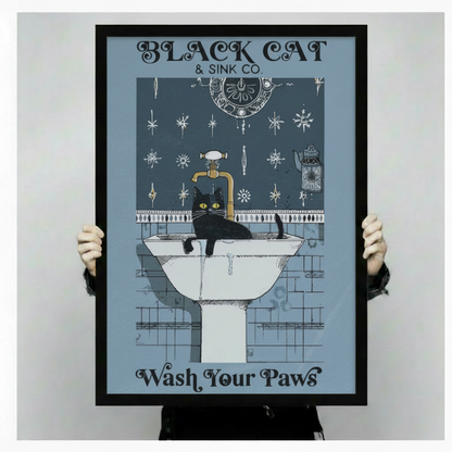 Hands holding large framed Black Cat and Sink Co Wash Your Paws canvas print - full artwork detail of vintage gothic typography with yellow-eyed black cat draped over white pedestal sink brass faucet ornate medallion and dark blue snowflake tile pattern on steel blue background