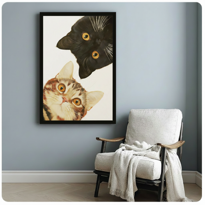 Black framed two cats peeking art print on blue-gray wall beside linen armchair, realistic cat portrait with amber eyes for minimalist home decor