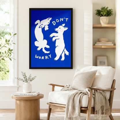 Large black framed pop art "Don't Worry" white dancing dogs on cobalt blue background beside a white upholstered wood-frame armchair with a linen throw blanket, round solid wood side table with a coffee table book, and open shelving with potted greenery