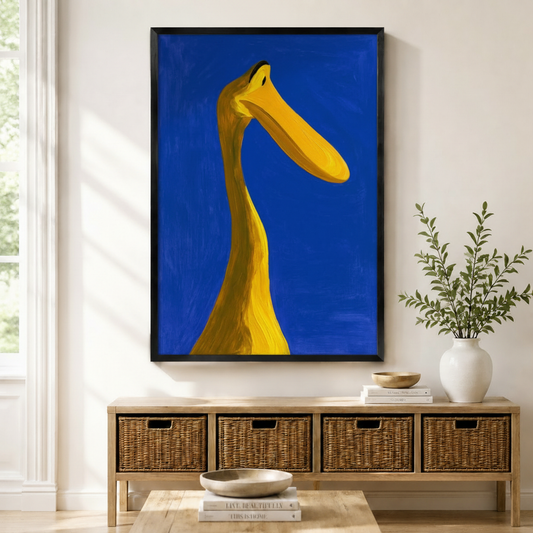 Black framed bold expressionist yellow duck neck close-up canvas art with a dramatic low-angle view of a long curved golden yellow bird neck stretching from the bottom to the top of the canvas with a wide flat beak extending horizontally to the right and a single small black eye on a rich cobalt blue impasto oil paint background, displayed above a wooden rattan storage bench with a white ceramic bowl, stacked books, and a white vase of greenery
