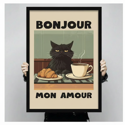 Close-up size reference of a black-framed retro poster print featuring a grumpy black cat at a café table with croissant and coffee and bold "Bonjour Mon Amour" lettering, held by two hands