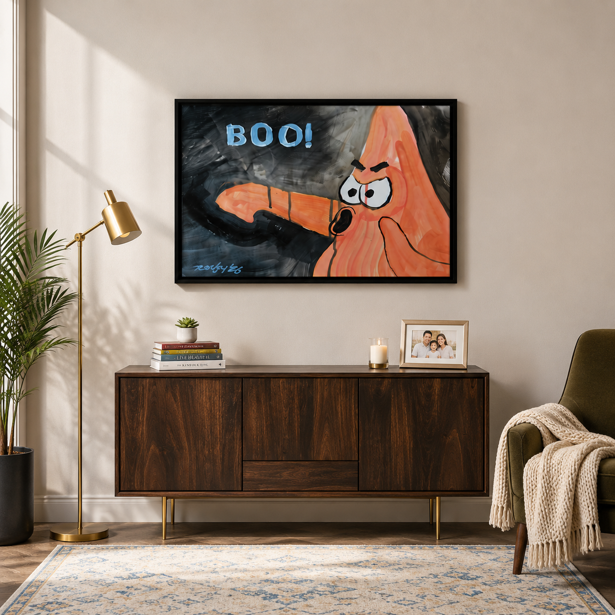 Framed bold cartoon BOO wall art print displayed above wooden sideboard in warm-toned living room with gold floor lamp and green armchair