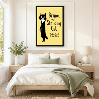 Black framed Bruno the Standing Cat whimsical book cover canvas art with a round-eyed black and white tuxedo cat standing upright with a startled expression beside bold title lettering and script author names Nadine Robert and Jean Jullien on a bright lemon yellow background, hung above a wooden bed frame in a sun-drenched Scandinavian bedroom with crisp white linen bedding, a sage green accent pillow, matching round nightstand lamps, and a white ceramic vase of greenery