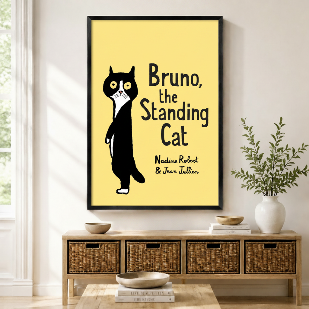 Black framed Bruno the Standing Cat whimsical book cover canvas art with a wide-eyed black and white tuxedo cat standing upright beside the bold hand-lettered title Bruno the Standing Cat and author names Nadine Robert and Jean Jullien on a bright lemon yellow background, displayed on a light wall above a wooden rattan storage bench with wooden bowls, stacked books, and a white ceramic vase of greenery