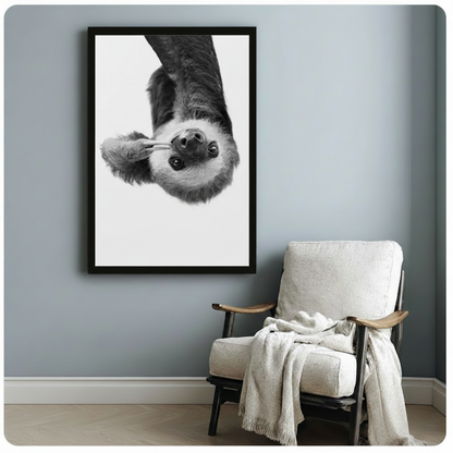 Black framed black and white realistic sloth hanging upside down staring forward with fluffy fur and peace sign claws canvas art on white background on a soft blue-gray wall beside a white linen folding armchair with a cream throw blanket
