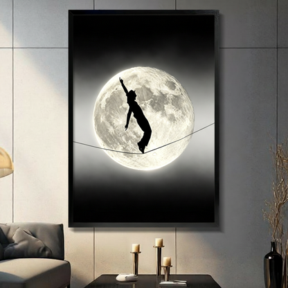 Black and white surreal photograph of a cat-girl silhouette with cat ears and tail balancing on a tightrope in front of a glowing full moon on a dark night sky wall art framed print in living room with light gray panel wall and dark sofa