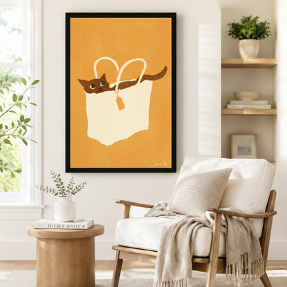 Large black-framed wall art of a laid-back brown cat sprawled inside a cream tote bag against a warm amber background in clean painterly style, styled beside a linen armchair in a Scandinavian-inspired reading nook