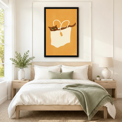 Oversized black-framed canvas of a relaxed brown cat nestled inside a cream shopping bag on a golden mustard background, mounted as a warm boho statement piece above a neutral linen bed in a serene minimalist bedroom