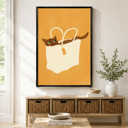 Black-framed canvas print of a whimsical brown cat lounging inside a cream tote bag on a warm mustard orange background, displayed above a rattan storage console in a bright Scandinavian entryway