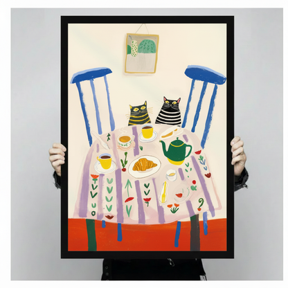 Person holding a black framed illustration of two striped cats at a tea party with a green teapot, croissant, and blue chairs against a light grey backdrop, full front view of whimsical animal kitchen wall art poster print