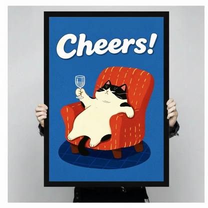 Close-up of black framed Cheers cat wall art print showing chubby tuxedo cat toasting with wine glass in red armchair on blue background, held by person