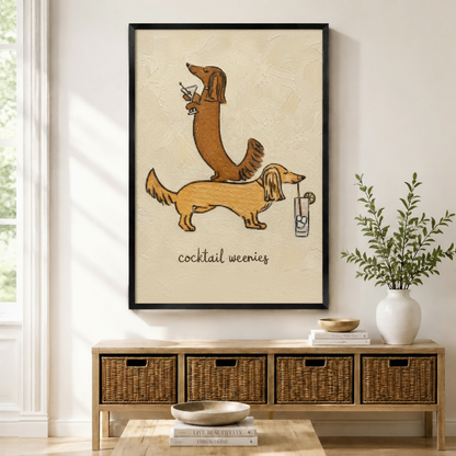 Black-framed canvas print of two whimsical long-haired dachshunds stacked together holding cocktail drinks with "cocktail weenies" lettering in textured oil painting style, displayed above a rattan storage console in a bright modern entryway
