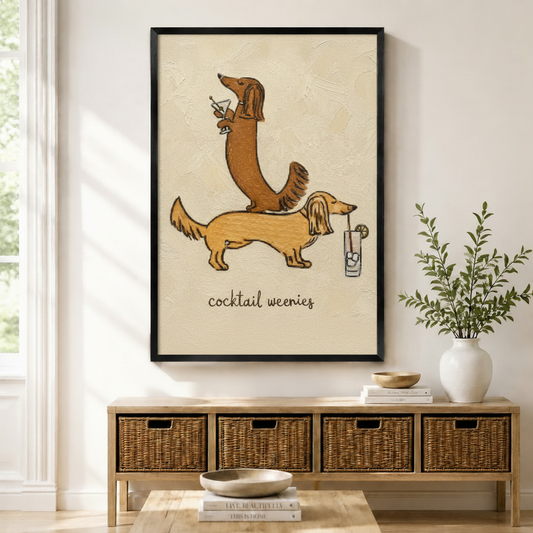 Black-framed canvas print of two whimsical long-haired dachshunds stacked together holding cocktail drinks with "cocktail weenies" lettering in textured oil painting style, displayed above a rattan storage console in a bright modern entryway