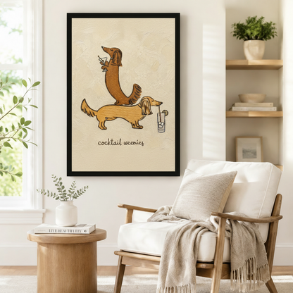 Large black-framed wall art of two long-haired dachshunds with cocktail glasses in expressive textured illustration style with "cocktail weenies" script, styled beside a linen armchair and round side table in a Scandinavian reading corner