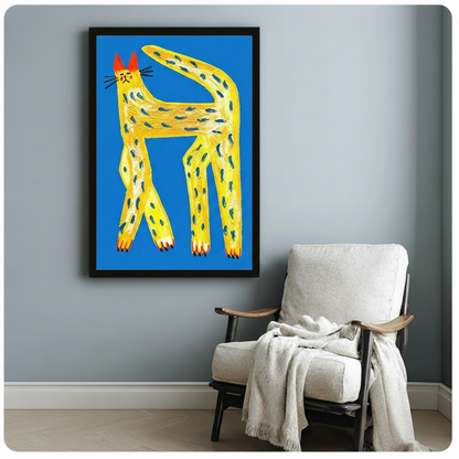 Black framed canvas of a whimsical yellow cat with teal spots and red-tipped ears on a cobalt blue background, mounted on a blue-grey wall beside a white linen armchair