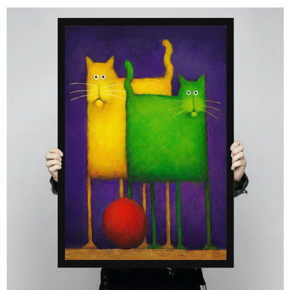 Person holding a large black framed bold colorful cat duo canvas art print showing a quirky tall yellow cat and a chunky green cat standing together with a bright red ball between their long legs on a vivid purple background against a light gray studio backdrop