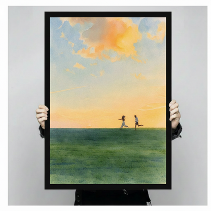 Framed canvas art print of couple running on green meadow under glowing golden sunset with soft peach clouds, held up showing full artwork detail and black frame