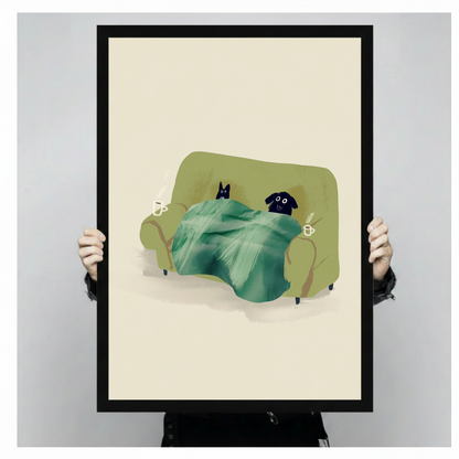 Person holding a large vertical black framed cozy cat and dog snuggled under a blanket canvas art print showing a black cat and black dog resting on an olive green sofa beneath a green blanket with two steaming coffee cups on a cream background in a light gray studio setting