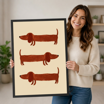 Smiling woman holding a large black-framed canvas print of three illustrated dachshunds, showing print size, frame quality, and artwork detail up close