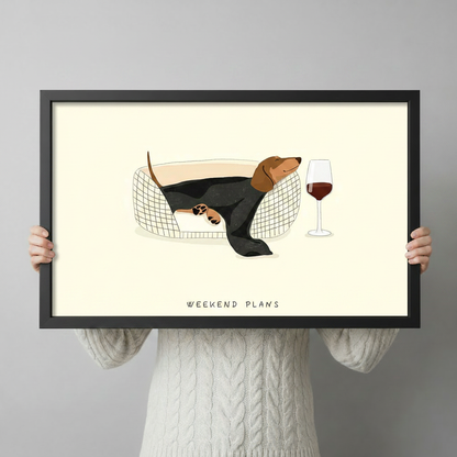 Person holding a black framed art print of a dachshund resting in a basket with a red wine glass, cream background with Weekend Plans text