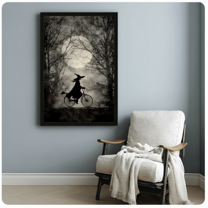 Black framed dark academia witch on bicycle moonlit forest silhouette canvas art in moody black and gray on a soft blue-gray wall beside a white linen lounge chair with a cream throw blanket draped over the arm