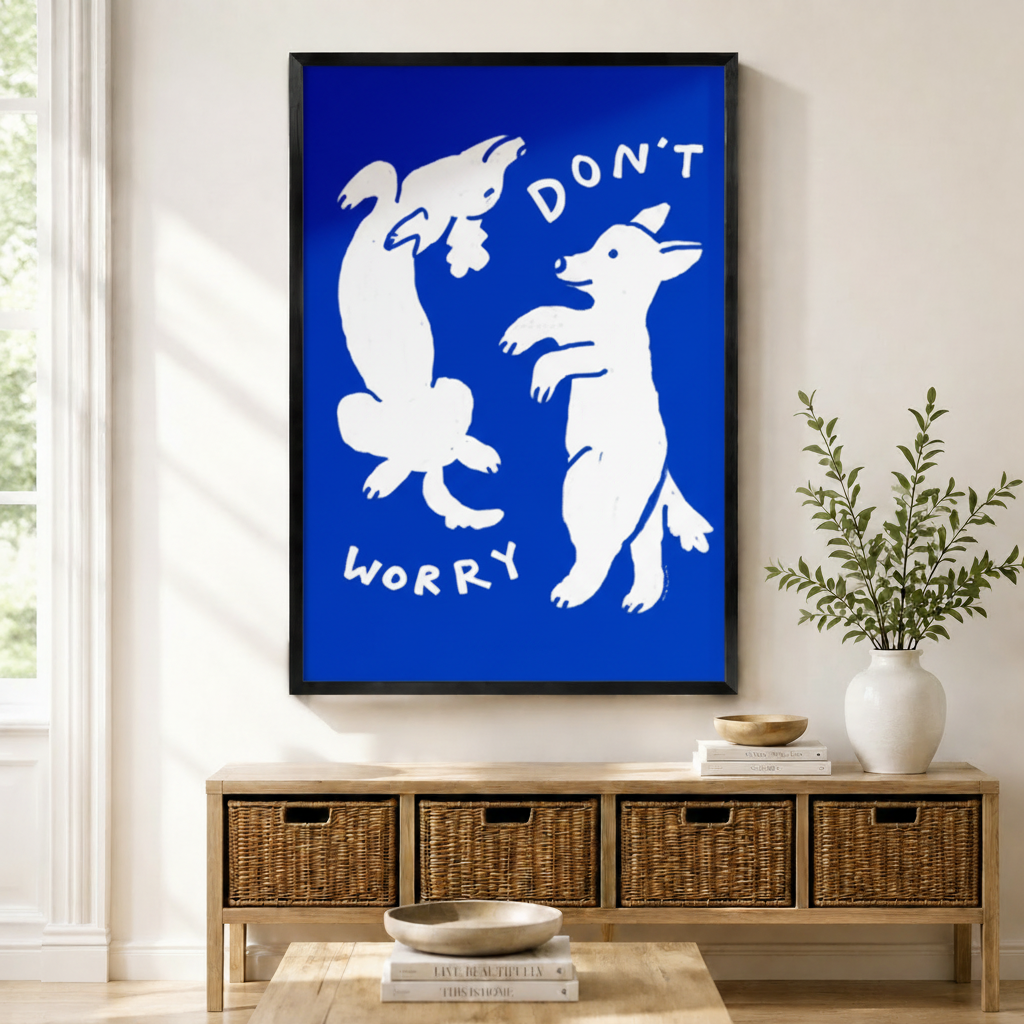 Black framed bold blue "Don't Worry" dancing dogs wall art with white line illustration mounted above a light wood wicker basket console with ceramic bowls, stacked books, and a tall white vase in a warm minimal entryway