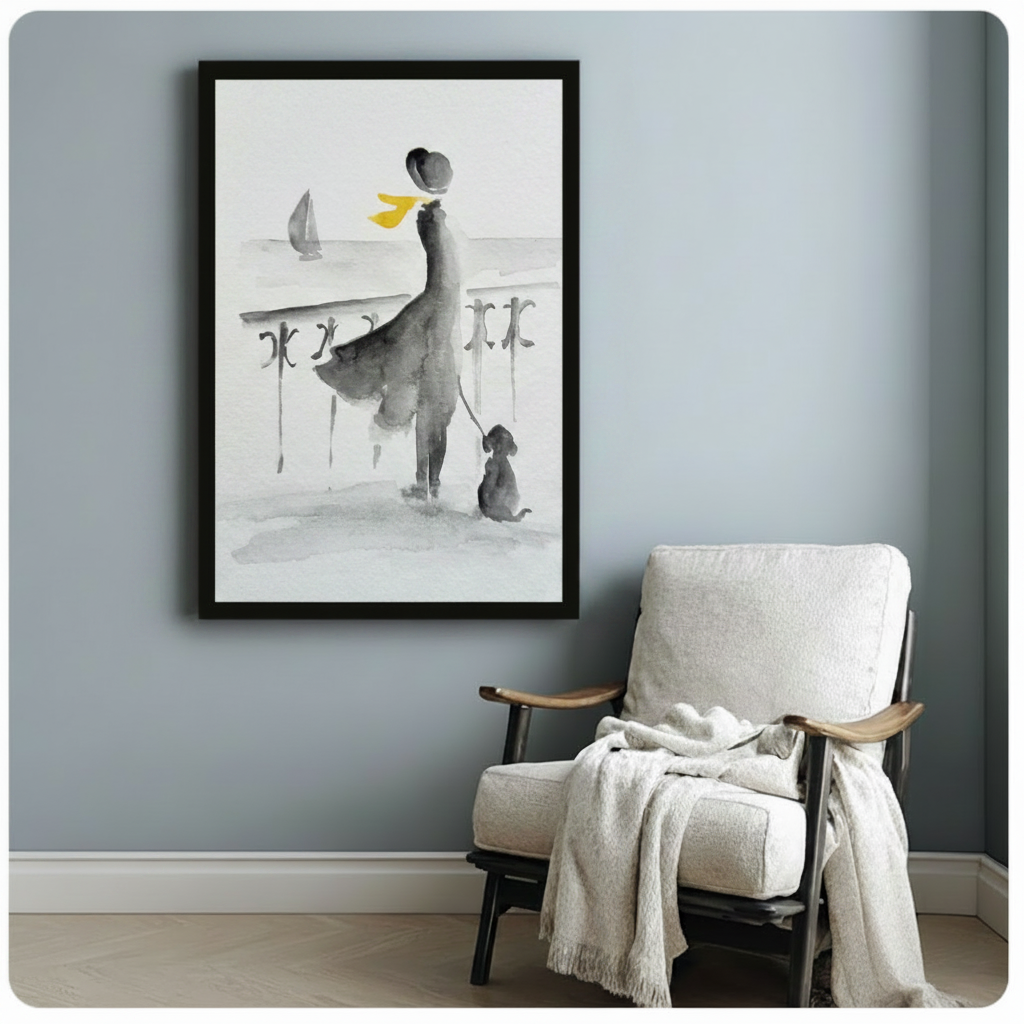 Black-framed watercolor wall art depicting a stylish woman and her dog overlooking the ocean, displayed on a blue-grey wall beside a white armchair