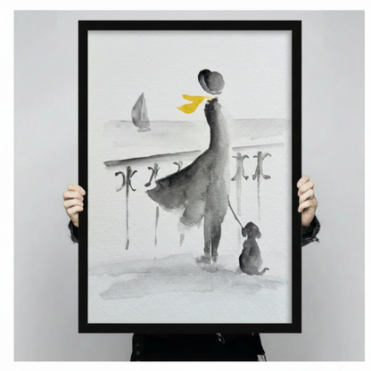Close-up hand-held view of a black-framed watercolor canvas print showing an elegant woman with yellow scarf and dog at a seaside railing