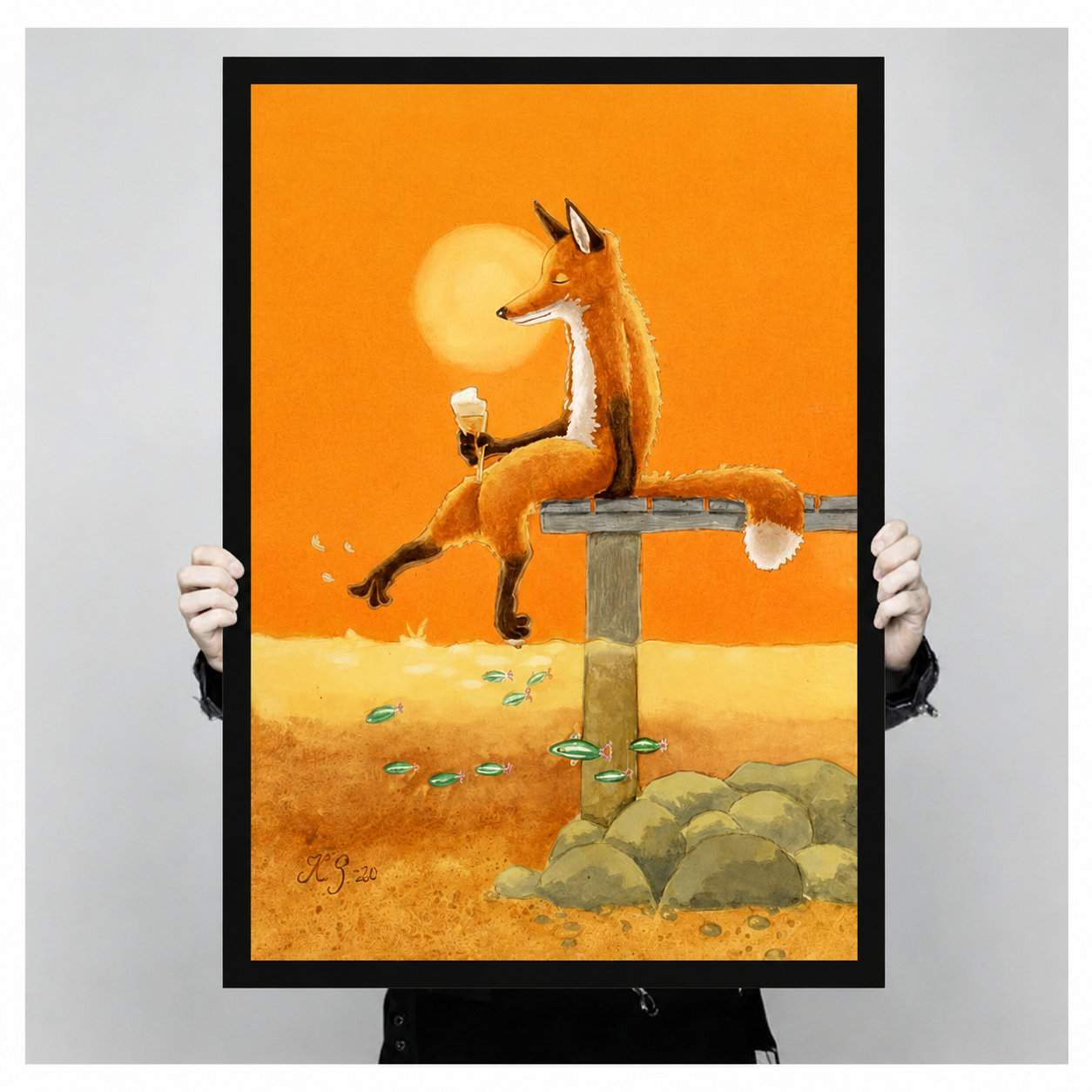 Close-up of framed poster showing fox sitting on bench holding wine glass with floating fish and orange sunset sky held by hands
