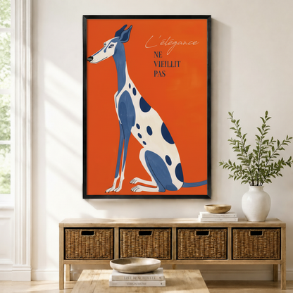 Black framed retro French poster greyhound canvas art with an elegant blue and white spotted greyhound sitting upright beside the script quote L'élégance ne vieillit pas on a vivid orange-red background, displayed on a light wall above a wooden rattan storage bench with a white ceramic vase of greenery and stacked books on the floor