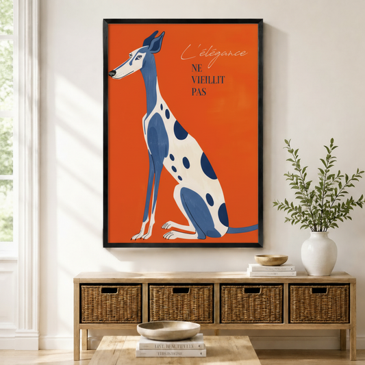 Black framed retro French poster greyhound canvas art with an elegant blue and white spotted greyhound sitting upright beside the script quote L'élégance ne vieillit pas on a vivid orange-red background, displayed on a light wall above a wooden rattan storage bench with a white ceramic vase of greenery and stacked books on the floor