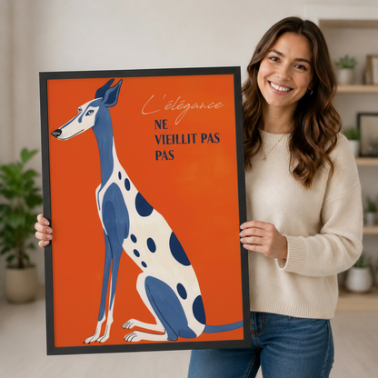 Smiling woman holding a large vertical black framed retro French greyhound canvas art print showing an elegant blue and white polka-dot greyhound seated upright with the French script quote L'élégance ne vieillit pas on a vivid orange-red background in a bright plant-filled room