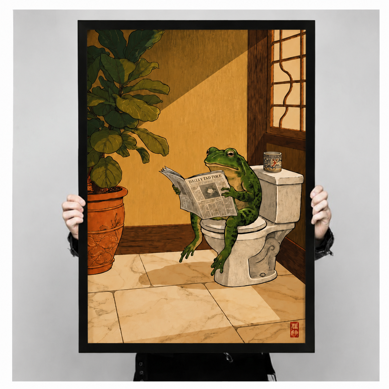 Person holding large black framed print of humorous frog reading newspaper on toilet, funny bathroom wall art gift idea