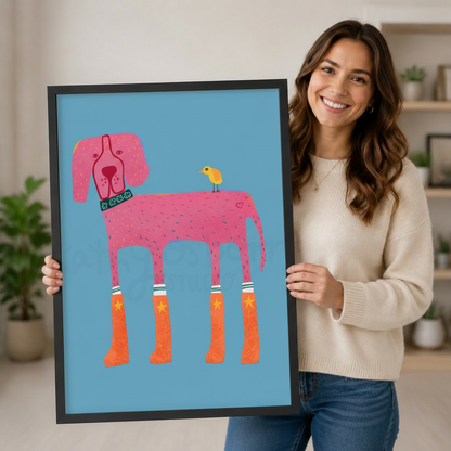 Smiling woman in cream sweater holding a black framed cartoon pink dog wearing orange star rain boots with a yellow bird art print in sky blue against a neutral indoor background with potted plants