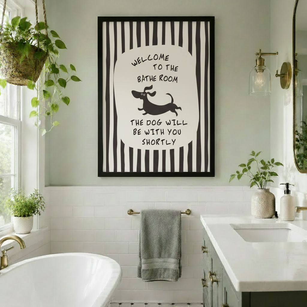 Black framed whimsical bathroom dachshund quote canvas art with bold black and white vertical stripes and a central oval with hand-lettered welcome to the bathroom above a bounding cartoon dachshund silhouette and the text the dog will be with you shortly hung in a bright bathroom with a freestanding white soaking tub, white subway tiles, gold hardware, a towel bar, hanging potted plants, and a double vanity