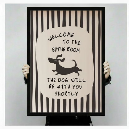 Person holding a large vertical black framed funny bathroom dachshund stripe canvas art print showing bold black and white vertical stripes with a central oval featuring hand-lettered welcome to the bathroom above a jumping cartoon dachshund silhouette and the humorous quote the dog will be with you shortly against a light gray background