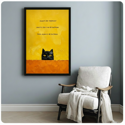 Black framed whimsical black cat peeking canvas art with a cheeky always be batman quote in bold lettering on a sunny yellow and orange textured background, displayed on a blue-gray wall beside a white linen armchair draped with a cream throw blanket