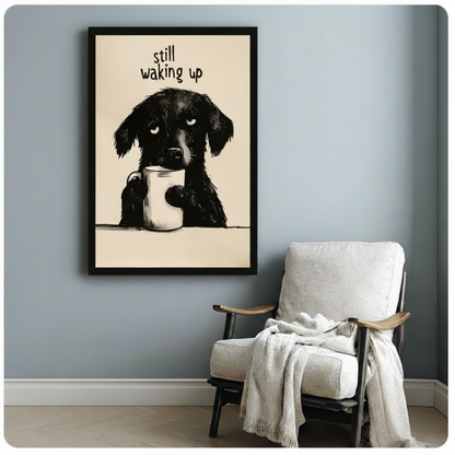 Black framed funny illustration sleepy black dog hugging white coffee mug "Still Waking Up" quote canvas art on warm beige background on a soft blue-gray wall beside a white linen folding armchair with a cream throw blanket