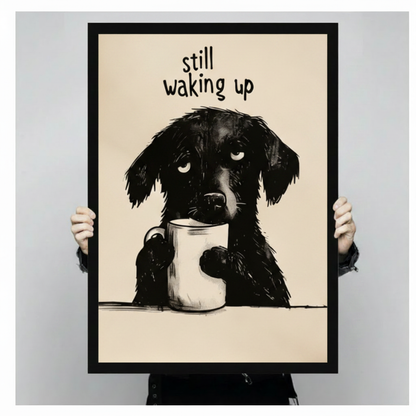 Person holding a black framed humorous pencil illustration drowsy black dog clutching white coffee mug "Still Waking Up" art print on warm beige background against a light gray backdrop