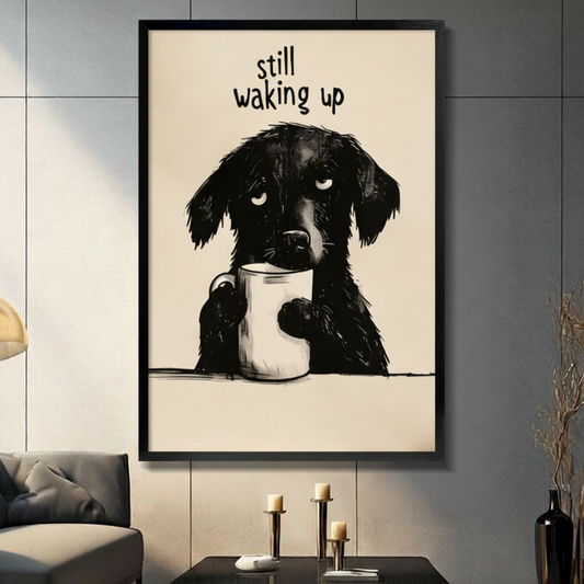 Funny illustration sleepy black dog holding white coffee mug with drowsy eyes "Still Waking Up" quote black framed canvas art on warm beige background on a dark gray paneled wall above a charcoal sofa with brass candle holders and a black decorative vase