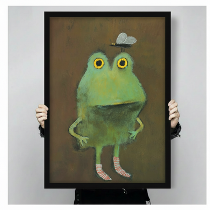 Close-up product shot of a framed wall art print showing a humorous green frog with a butterfly on its head and striped socks, earthy brown background with painterly texture